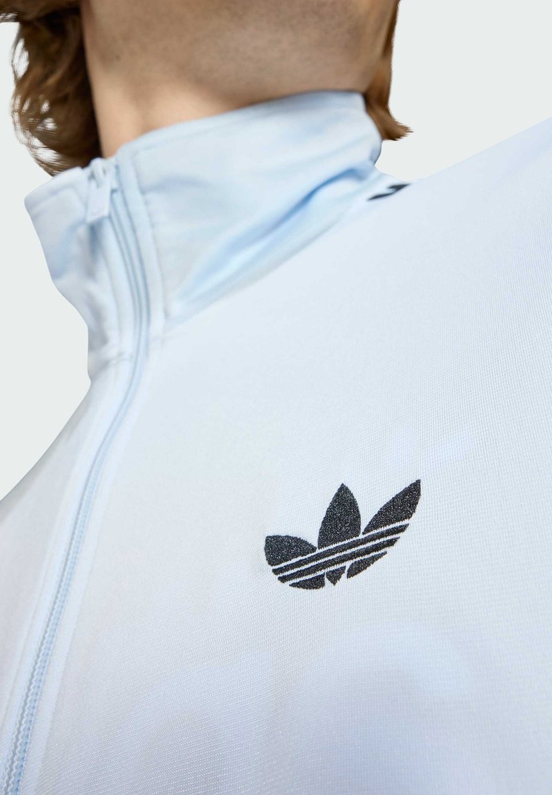 Persona che indossa una giacca con zip azzurro chiaro con il logo Adidas Trefoil nero sul petto sinistro, visibile dal collo alla parte superiore del busto.