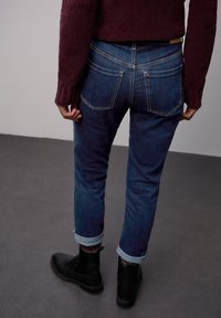 Pantalones vaqueros de mezclilla azul oscuro con corte de pierna recta, bolsillos traseros visibles y dobladillos remangados. Combinados con botines negros y un suéter burdeos.