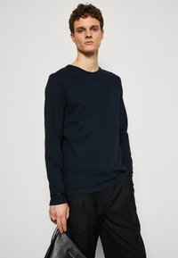 Camiseta de manga larga en color navy con cuello redondo. Hecha de algodón, presenta un pequeño parche de logo en el pecho. Combinada con pantalones negros y un bolso.