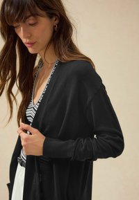 Cardigan nero con una texture morbida, design aperto davanti e polsini a coste. Indossato sopra una maglia a righe bianco e nero.