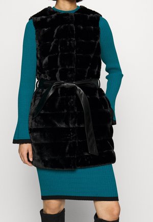 Svart fuskpälsväst med en texturerad, quiltad design, som har ett knutet svart bälte. Bärs över en blå ribbad långärmad klänning.