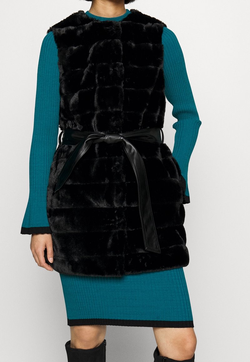 Svart fuskpälsväst med en texturerad, quiltad design, som har ett knutet svart bälte. Bärs över en blå ribbad långärmad klänning.