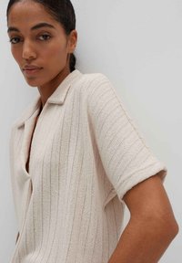 Helles beiges Kurzarm-Strickshirt mit kragenförmigem Halsausschnitt, vertikal gerippter Textur und lockerer Passform. Keine sichtbaren Verschlüsse.