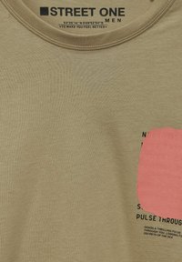 Camiseta de manga corta color caqui con cuello redondo; presenta un acento texturizado en rosa y texto impreso en negro en el lado izquierdo.