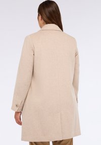 Abrigo de mezcla de lana beige con corte recto, gran solapa y puños abotonados, que presenta una abertura en la parte posterior y un diseño con detalles mínimos.