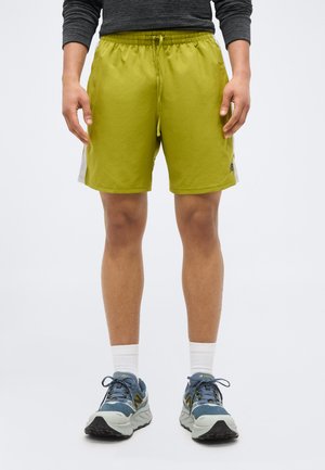 Άτομο που φοράει αθλητικά σορτς λάιμ πράσινου χρώματος, λευκές κάλτσες και μπλε-γκρι παπούτσια τρεξίματος, στέκεται με τα χέρια χαλαρά δίπλα στο σώμα.