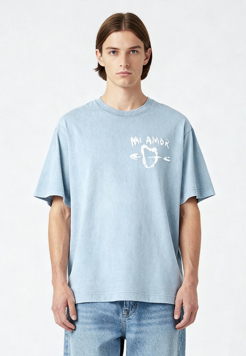 T-shirt in cotone azzurro chiaro con vestibilità rilassata, colletto rotondo e maniche corte, caratterizzata da una grafica bianca "Mi Amor" sul davanti.