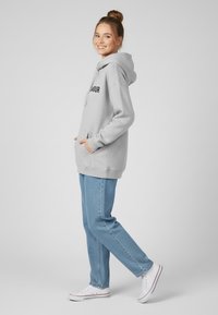 Felpa grigia con grande tasca frontale e testo nero, abbinata a jeans blu chiaro a gamba dritta e sneakers bianche, il tutto su uno sfondo semplice.