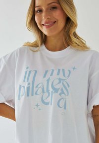 Biały, oversize'owy t-shirt z jasnoniebieskim napisem "w mojej erze pilates" w stylizowanej czcionce z drobnymi gwiazdkami. Miękka tkanina, luźny krój.