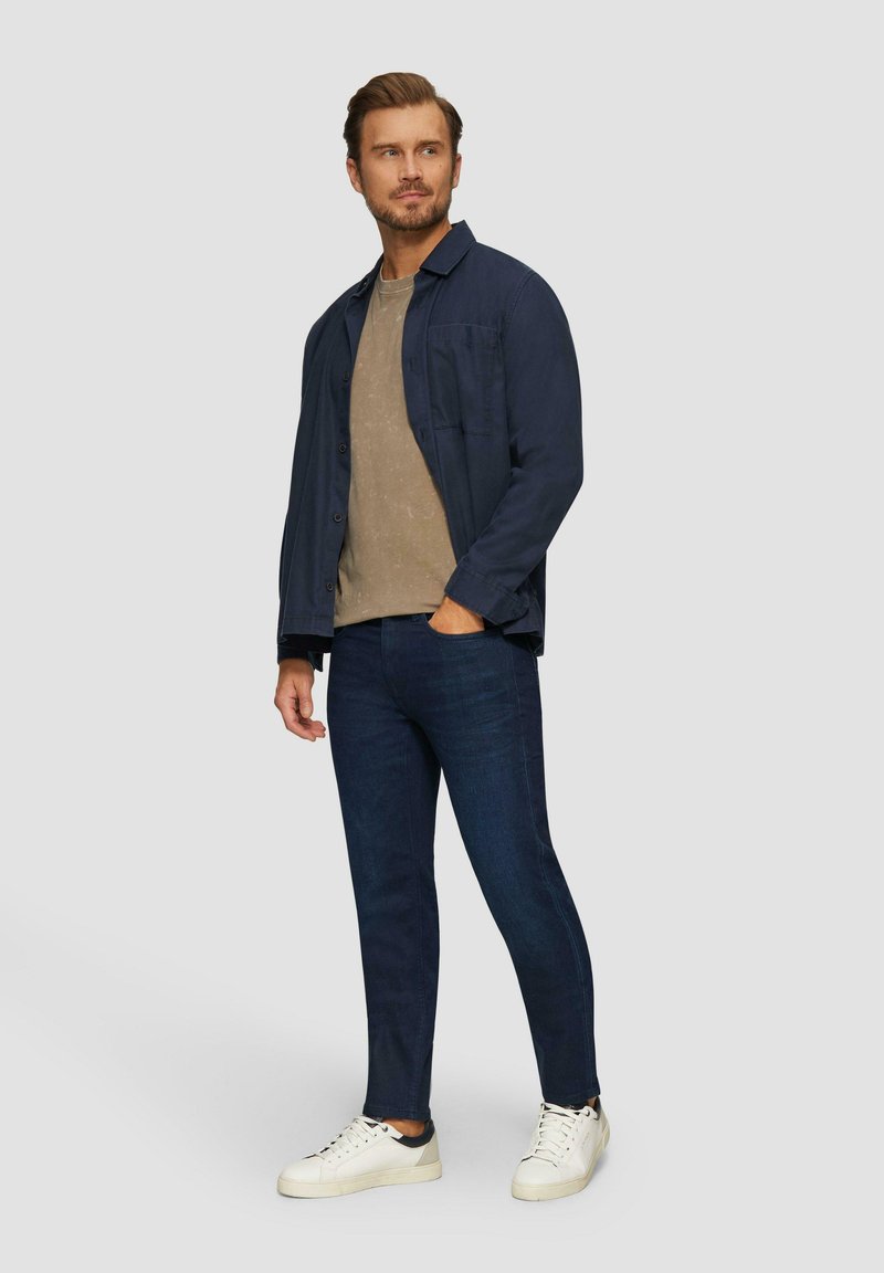 Chemise bleu marine à boutons portée sur un t-shirt beige clair, associée à un jean foncé et des baskets blanches, mettant en avant un look décontracté en couches.