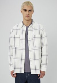 Chemise à carreaux blanche et violette en tissu texturé, dotée de deux poches à la poitrine, d'une fermeture à boutons et d'une coupe décontractée. Portée par-dessus une chemise grise.