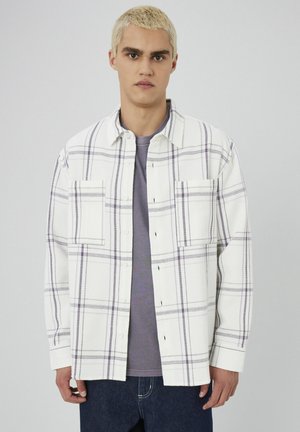 Chemise à carreaux blanche et violette en tissu texturé, dotée de deux poches à la poitrine, d'une fermeture à boutons et d'une coupe décontractée. Portée par-dessus une chemise grise.