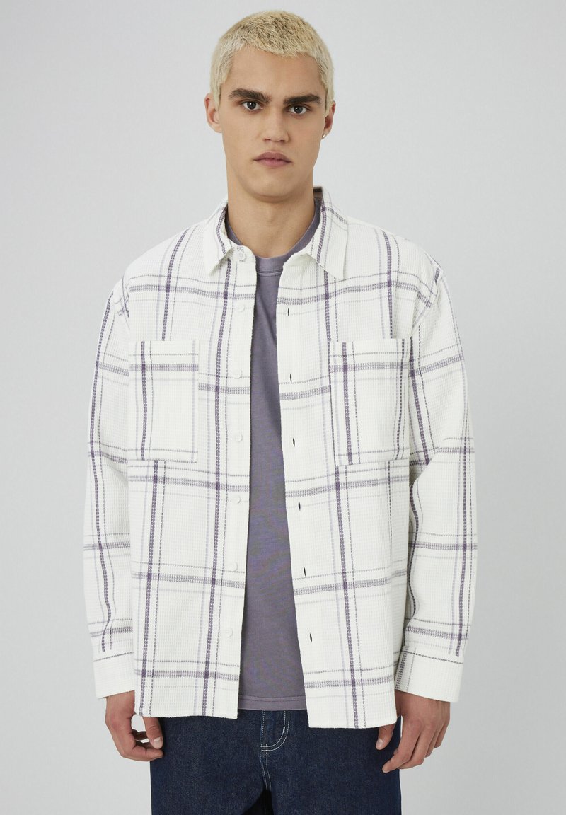 Chemise à carreaux blanche et violette en tissu texturé, dotée de deux poches à la poitrine, d'une fermeture à boutons et d'une coupe décontractée. Portée par-dessus une chemise grise.