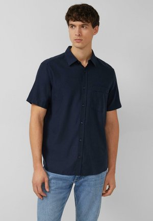 Jeune homme portant une chemise bleu marine à manches courtes boutonnée et un jean bleu clair, regardant légèrement vers sa gauche sur un fond uni.