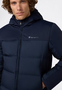 Giacca a vento imbottita blu navy con cappuccio, dotata di chiusura a zip e texture trapuntata. Logo "Champion" argento sul petto.