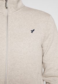Chaqueta beige con cremallera y cuello alto, con pequeño logotipo de pájaro bordado en oscuro en el lado izquierdo del pecho.
