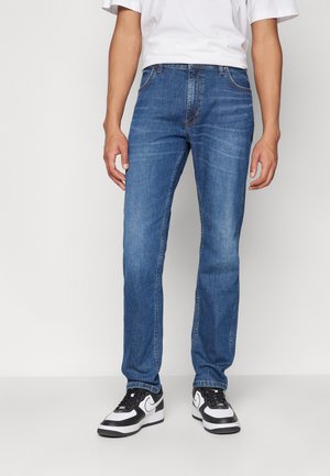 Modre kavbojke s ravnim krojem, opremljene s petimi žepi, diskretnimi šivi in obrabljenim videzom, v kombinaciji s črno-belimi supergami.