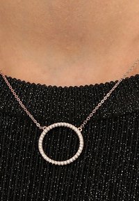 Collana in oro rosa con un pendente circolare, incastonato di gemme trasparenti, pende da una delicata catena su un maglione nero a coste.