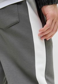 Sive trenerke s bijelim bočnim trakama, napravljene od mekog materijala. Imaju stražnji dzep i glatku teksturu. Ruka počiva na tkanini.
