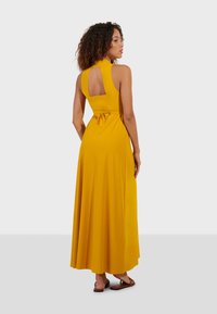 Dlouhé žluté šaty s halter výstřihem, odhaleným zádům a detailem v pase na stahování. Hladká tkanina elegantně splývá až k zemi.