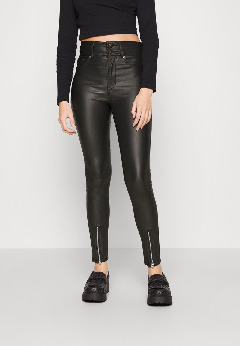 Femme portant un pantalon noir en similicuir taille haute avec poches avant et fermetures éclairs aux chevilles, associé à des mocassins noirs épais et un top court à manches longues.