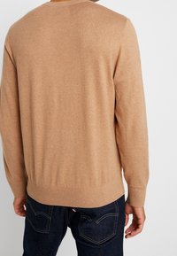 Maglione beige a maglia con colletto rotondo, polsini e orlo inferiore a coste, visto da dietro, abbinato a jeans in denim scuro.