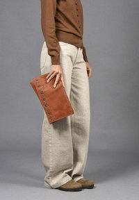 Clutch de cuero marrón con accents de tachuelas plateadas, sostenido en una mano. Combinado con pantalones de pierna ancha beige de cintura alta y un cárdigan marrón.