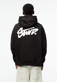 Černá mikina s kapucí a velkým bílým nápisem na zádech. Má šňůrkovou kapuci a žebrované manžety. Nosená se světlými kalhotami.