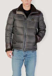 Uomo che indossa un giubbotto imbottito grigio scuro con colletto e polsini in shearling marrone, abbinato a jeans azzurri chiari, in piedi contro uno sfondo bianco.