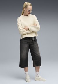 Sudadera blanca corta con puños acanalados, jeans oscuros de pierna ancha, zapatillas blancas y calcetines cortos blancos con un logo. Fondo gris.