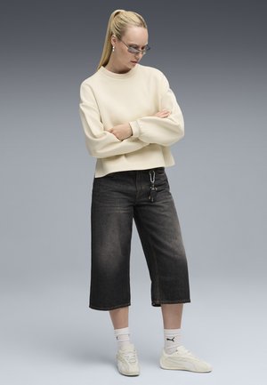 Felpa bianca corta con polsini a costine, jeans neri a gamba larga, sneakers bianche e calzini bianchi corti con logo. Sfondo grigio.