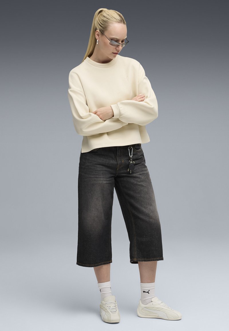 Sudadera blanca corta con puños acanalados, jeans oscuros de pierna ancha, zapatillas blancas y calcetines cortos blancos con un logo. Fondo gris.