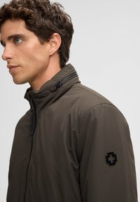 Olivgrüne Jacke mit hohem Kragen, Reißverschluss und schwarchem Logo-Patch auf der Schulter; glatte Textur und leichtes Material.