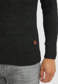 Svart ribbestrikket genser med en teksturert finish, med et beige logo-patch nederst og tettsittende mansjetter. Brukt sammen med lysegrå jeans.