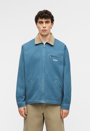 Ung mand iført en blå lynlåstrøje med beige krave og beige bukser, stående foran en ensfarvet lysegrå baggrund.