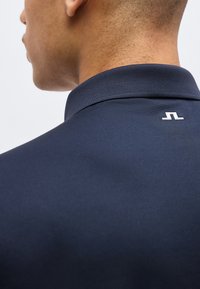 Κοντινό σε λαιμό και ώμο ατόμου που φοράει μπλε πουκάμισο με γιακά και μικρό λευκό λογότυπο στο πίσω μέρος του γιακά.