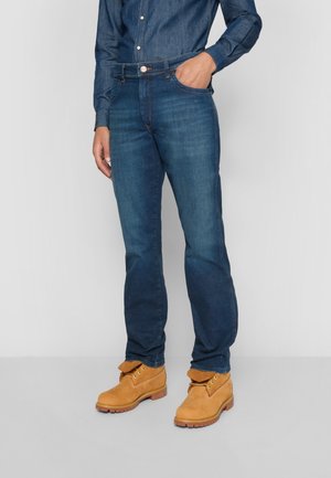 Person wearing blue denim jeans, a matching blue denim shirt, and tan lace-up work boots, standing with one hand in pocket.