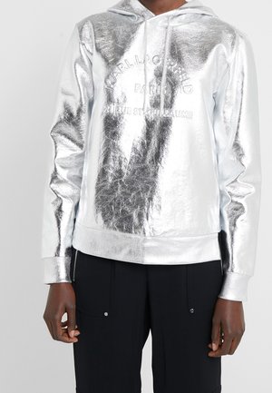 Henkilö, joka pukeutunut kiiltävään hopeanväriseen metallinhohtoiseen huppariin, jossa on etuosassa kohokuvioitu teksti, ja mustiin housuihin, seisoo yksinkertaista vaaleaa taustaa vasten.