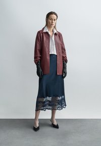 Bordowa skórzana kurtka na białej koszuli w paski, zestawiona z granatową spódnicą z przezroczystym koronkowym wykończeniem i czarnymi szpilkami. Czarnych rękawiczek dopełnia wygląd.