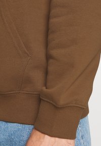 Gros plan sur le tissu d'un sweat-shirt marron, avec des poignets côtelés et un détail de poche latérale. Texture lisse avec des accents de couture visibles.