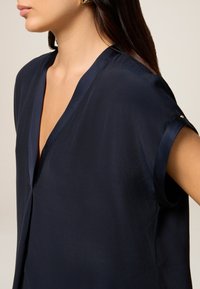 Blouse bleu marine avec un décolleté en V profond, des manches courtes et des détails en satin au niveau du col et des poignets ; texture lisse et design simple.