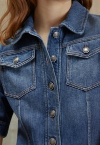 Camisa de denim con mangas cortas, cierre de botones en el frente, dos bolsillos en el pecho, color azul claro, detalles de costura visibles y botones de metal.