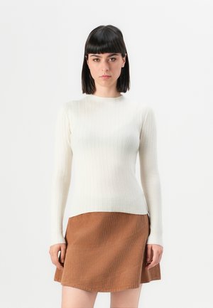 Femme aux cheveux noirs raides portant un pull à manches longues côtelé crème et une jupe en velours côtelé marron, se tenant devant un fond blanc uni.