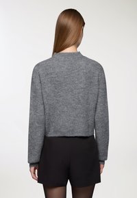 Kobieta z długimi prostymi włosami, ubrana w szary sweter dzianinowy i czarne szorty, widoczna z tyłu na jasnym, jednolitym tle.