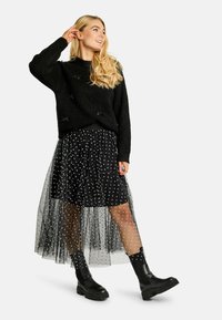 Čierny pletený sveter, navrstvený na čiernej tulejovej sukni s bodkami a striebornými srdcovými ozdobami. Čierne členkové čižmy dopĺňajú outfit.