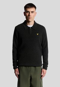 Ciemnoszara polo z długim rękawem, z kołnierzykiem, plisą na guziki oraz żółtym logo na lewej piersi, w połączeniu z zielonymi szortami typu cargo.