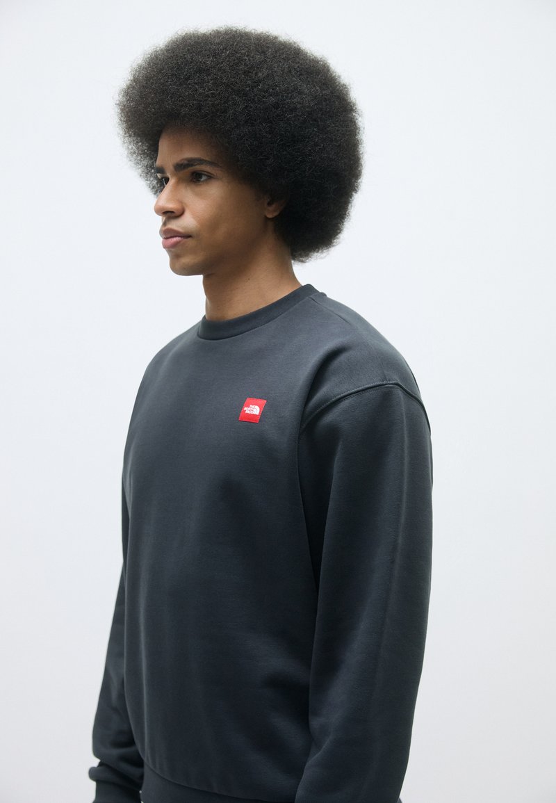 Sweatshirt gris foncé avec un col rond et des manches longues, arborant un petit patch logo rouge sur la poitrine. Texture lisse, coupe décontractée.