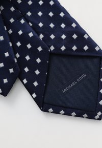 Cravate en soie bleu marine ornée d'un motif géométrique de petits diamants blancs. Doublure unie bleu foncé avec une étiquette en relief "MICHAEL KORS".