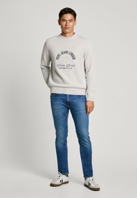 Hellgrauer, gestrickter Pullover mit Rundhalsausschnitt und Logo, kombiniert mit blauen Jeans und weißen Sneakers mit schwarzen Streifen.