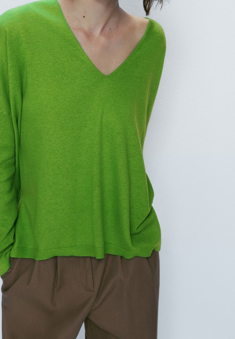 Personne portant un pull vert vif à col en V avec manches longues et un pantalon marron, les mains dans les poches, sur un fond clair uni.